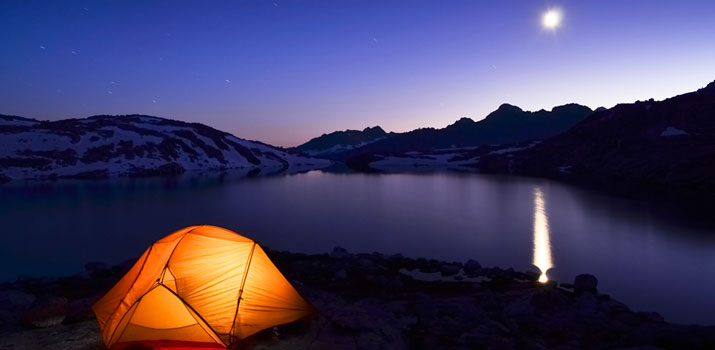 Tent lit up at night with a full moon beside a lake